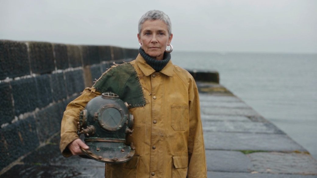 A woman in a mustard work jacket holds a brass diving helmet on a sea wall